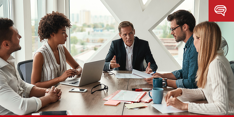 A group of five people are having a serious discussion around a conference table in a modern, well-lit office. One person, in the center, is presenting a pitch deck while others listen attentively. Laptops, documents, and coffee cups are on the table. Large windows provide a city view.