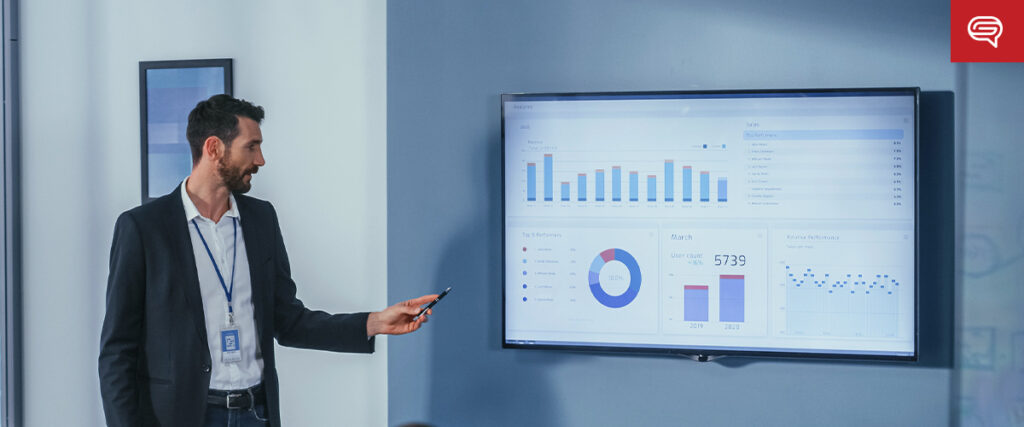 A man in a business suit stands next to a large screen displaying various charts and graphs. He is holding a pointer and gesturing towards the screen while presenting data from his pitch deck. The background features framed pictures and a light blue wall.