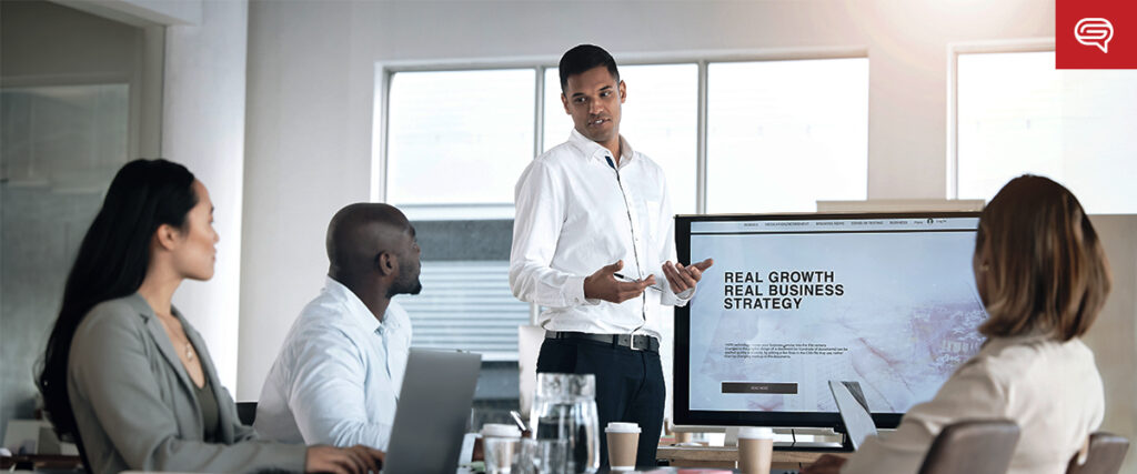 A business meeting with four people in a modern office. Three seated individuals are listening attentively to a standing man presenting information on a large screen. The screen reads "Real Growth Real Business Strategy." Various laptops and documents, along with an organized pitch deck, are on the table.