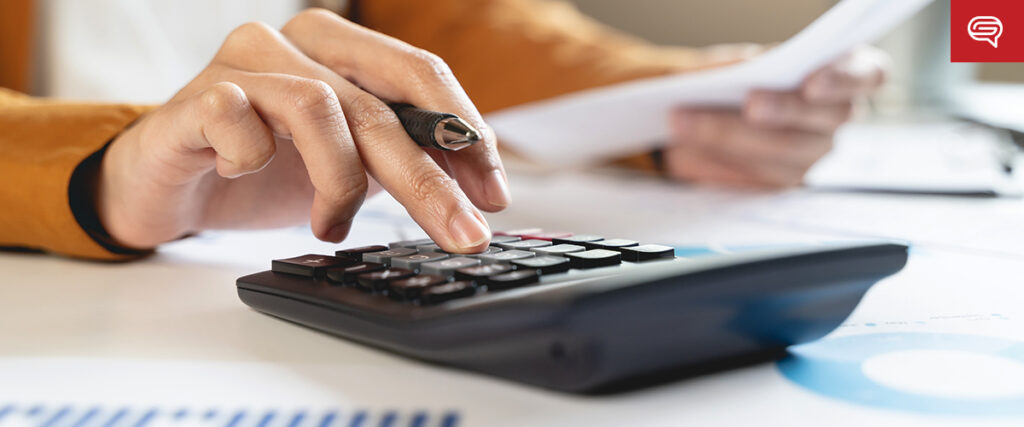 A close-up of a person using a black calculator while holding a pen and a piece of paper. The person is wearing an orange jacket. The background shows scattered documents and blurred office elements, indicating preparation for a detailed presentation.