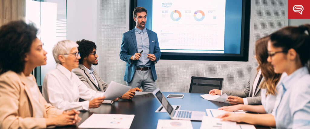 A team of six people sit around a conference table, looking at a standing man presenting a monthly budget report on a slide. The slide displays various colorful charts and graphs. The attendees are holding documents and appear engaged in the presentation.