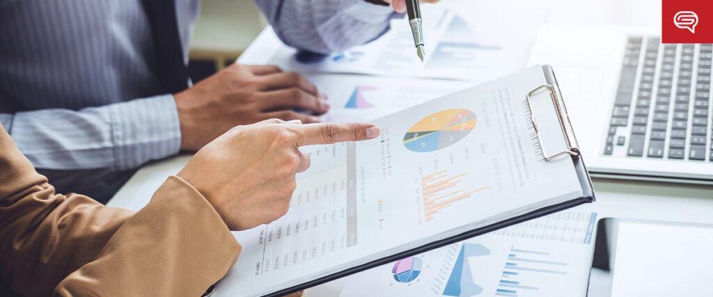 Two people at a desk review a clipboard with colorful pie and bar charts. One person points at a section of the chart while the other holds a pen and gestures toward the clipboard. A laptop and additional presentation materials are visible in the background.