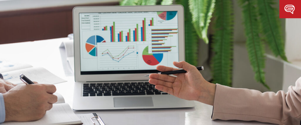 Two people are having a discussion, pointing at charts and graphs displayed on a laptop screen. The data includes bar graphs, pie charts, and line charts. They appear to be reviewing a PowerPoint presentation. Notebooks and pens are scattered on the table, with a plant in the background.