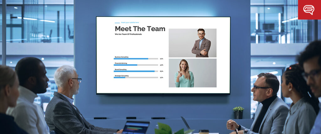 A team meeting in a modern office with people seated around a table looking at a large screen displaying a "Meet The Team" slide. The presentation shows photos of two individuals and a bar chart with percentages for different consulting specialties.