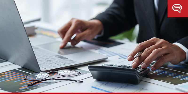 A person in business attire working on a laptop with one hand and using a calculator with the other. The workspace is covered with various charts and graphs, indicating preparation for a pitch deck. Glasses rest on the papers, and the red company logo is visible in the top right corner.