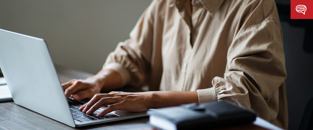 A person wearing a beige shirt types on a silver laptop at a wooden desk, seemingly preparing a presentation. An office chair and a closed black notebook are also visible on the desk. The background is blurred, focusing on the person’s hands and the laptop.