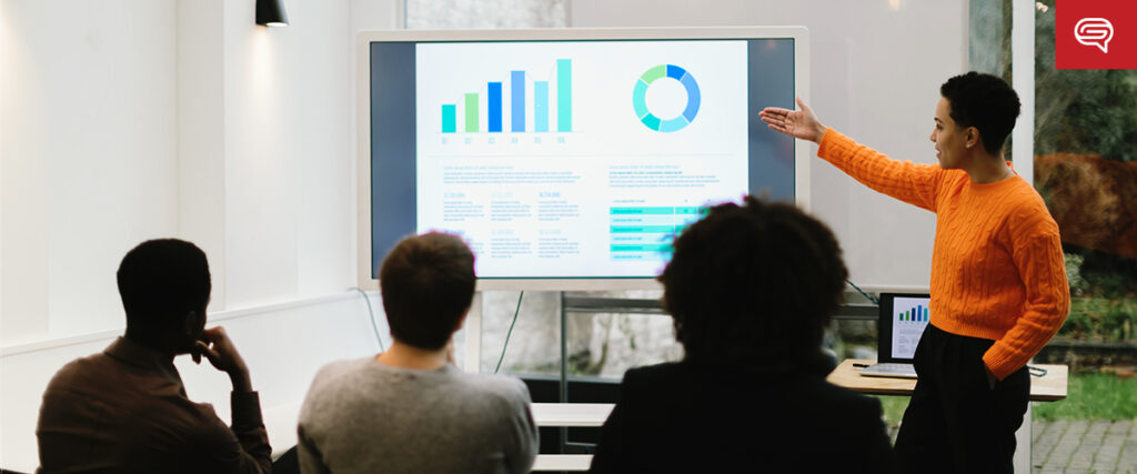 A person in an orange sweater is presenting data on a large screen to a group of people in a modern conference room. The screen displays various charts and graphs from the pitch deck. The audience members, seated and focused, watch the presentation attentively.