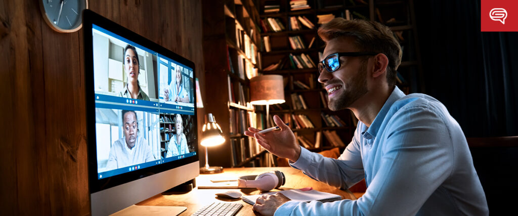 A man in glasses is sitting at a desk with a computer screen showing a video conference with four other people. He is smiling, gesturing with his hands, and appears engaged in the conversation about their PowerPoint presentation. The background includes a wooden wall and a bookshelf.