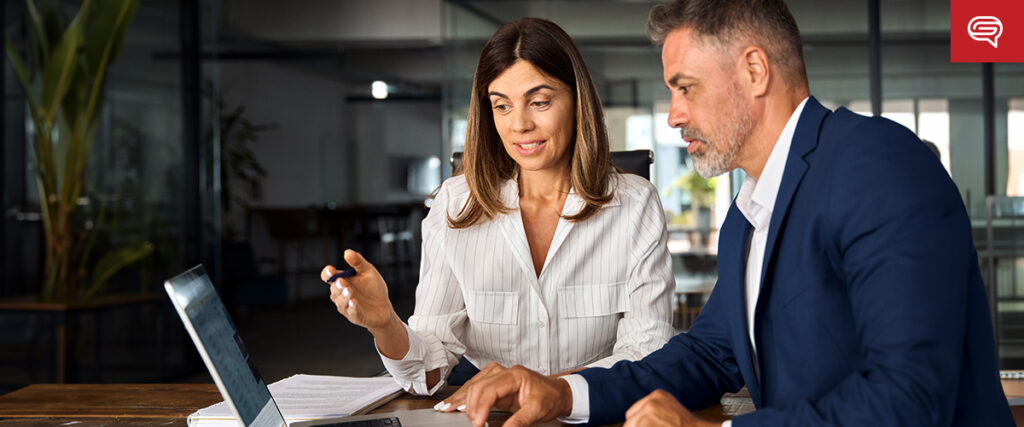 A woman and a man are sitting at a table in an office. The woman, holding a pen, is pointing at the laptop screen in front of them, while the man looks at the slide attentively. Both are dressed in business attire.