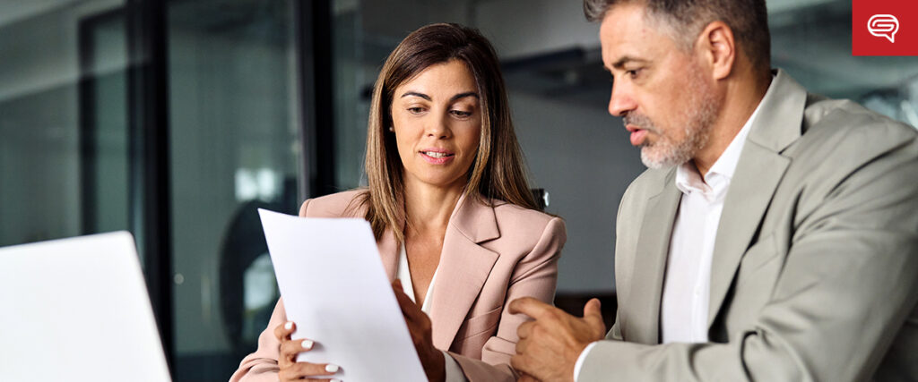 Two professionals, a woman and a man, are seated at a table in an office. They are both focused on a pitch deck that the woman is holding. The man is attentively gesturing towards the document while the woman reviews its content. A laptop is open on the table.
