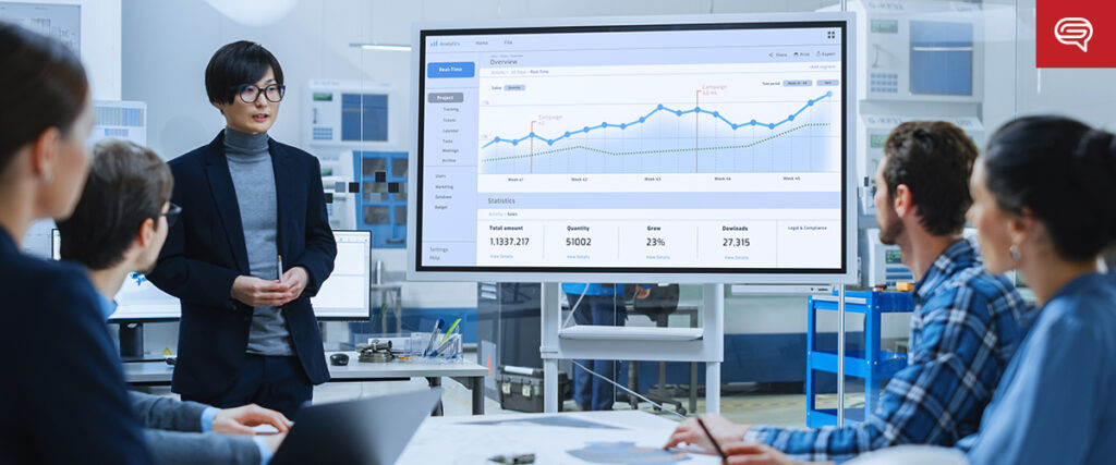 A person is presenting data on a large screen to a group of four people in a modern office setting. The screen displays PowerPoint slides with graphs and statistics, and all attendees appear focused on the presentation. The environment is bright with various computer equipment in the background.