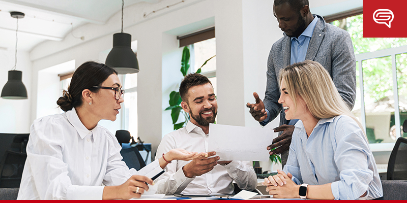 Four colleagues are gathered around a table in a modern office. Two men and two women are smiling and discussing a pitch deck. One woman is holding a pen and paper, while the others look on. The atmosphere appears positive and collaborative.