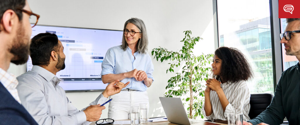 A group of five people are having a meeting in a modern office. One woman is standing and smiling while using a slide template for her presentation, and the others are seated, listening attentively. A large screen displaying a schedule is on the wall in the background, and a tall plant is nearby.