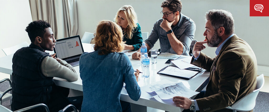 Five individuals are seated at a table in a conference room, engaged in a discussion. One person is using a laptop with a slide template open, and various documents and papers are spread across the table. Everyone appears focused and involved in the conversation.