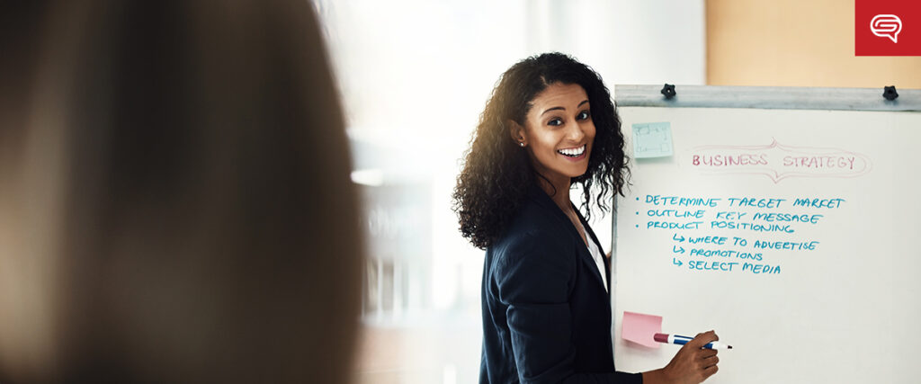 A woman with curly hair in a blazer is smiling while giving a presentation at a whiteboard. The whiteboard has a list titled "Business Strategy" with points such as "Determine Target Market" and "Select Media." Another person is blurred in the foreground.