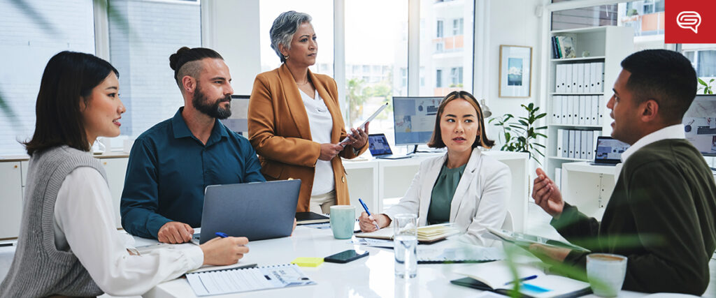 A diverse group of five people engaged in a business meeting in a modern office. Three are seated at a table with laptops and notebooks, while one stands with a tablet, listening attentively. Business charts and PowerPoint slides are visible on computer screens in the background.