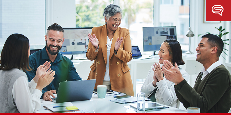 A diverse group of five coworkers sit around a table, clapping and smiling at a middle-aged woman among them, who is also clapping and smiling. Various electronic devices and documents on the table suggest they just witnessed an impressive slide presentation in their bright, modern office space.