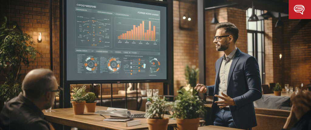 A man in a suit presents data visualizations on a large screen in a modern office space with brick walls. Potted plants and laptops are on the table, and two audience members listen attentively as he goes through the slide. The screen shows various charts and bar graphs from the pitch deck.