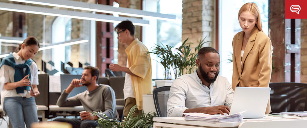 A modern office setting with five people engaged in various activities. Two people are having a conversation while one person is drinking coffee. A man is working on a laptop, preparing a pitch deck, and a woman is standing beside him. The office has large windows and plants.