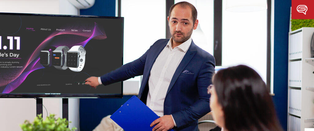 A man in a blue suit speaks while pointing at a screen displaying smartwatches and a promotional graphic. He holds a blue folder and stands in a modern office, with a woman seated, slightly out of focus, in the foreground, facing him. The presentation appears to be utilizing an effective slide template.