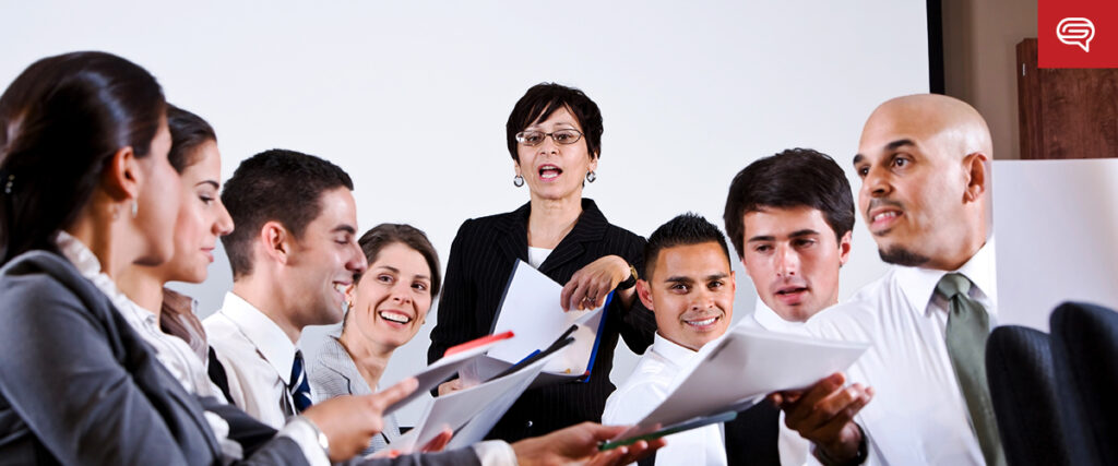 A group of business professionals sits and stands around a conference table in a meeting room, holding documents and laptops. A woman is standing in the center, gesturing as she presents a pitch deck. Everyone appears engaged in discussion.