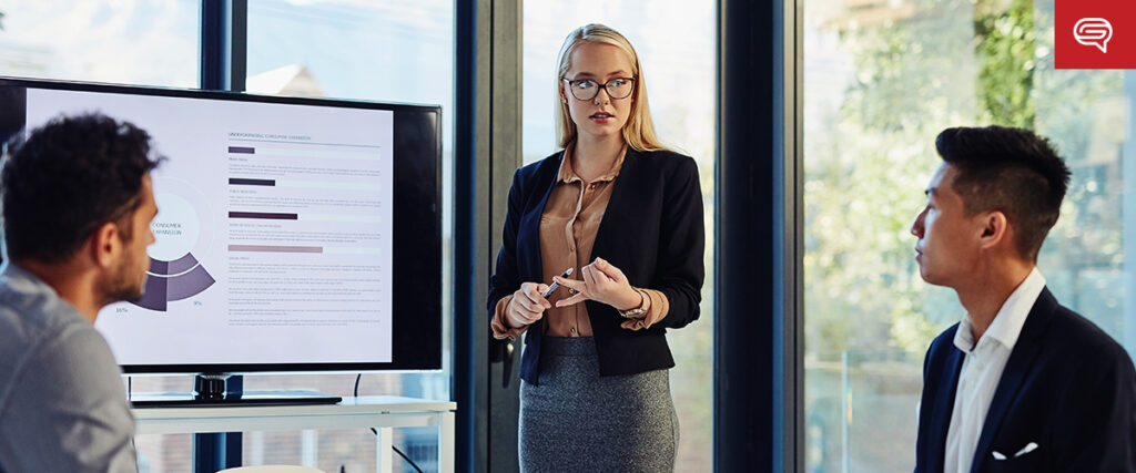 A woman in business attire stands and presents a pitch deck to two seated colleagues in an office setting. She uses a television screen displaying a chart with text and graphics. The room has large windows with outside greenery visible. A red logo is in the top right corner.