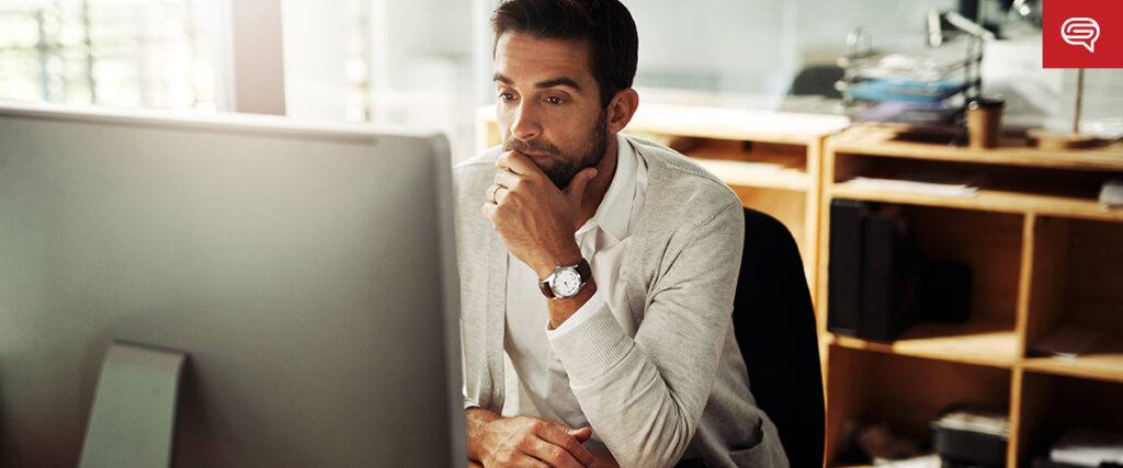 A man with a beard sits at a desk in an office, looking intently at a large computer monitor displaying a PowerPoint slide. He rests his chin on his hand, wearing a light-colored sweater and white shirt. Shelves with documents and office supplies are in the background.