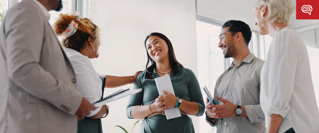 A diverse group of five people in a well-lit office setting are engaged in a positive conversation. One person holds documents, and another holds a notebook, likely discussing elements of a slide template. They appear happy and engaged, with smiles and gestures indicating lively interaction.