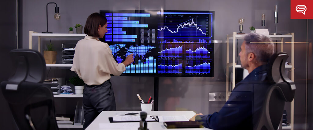 A woman in a professional setting points at a digital screen filled with various graphs, charts, and data visualizations from a pitch deck. A seated man watches her. The room features shelves with office supplies and a modern design. A coffee cup and devices are on the desk.