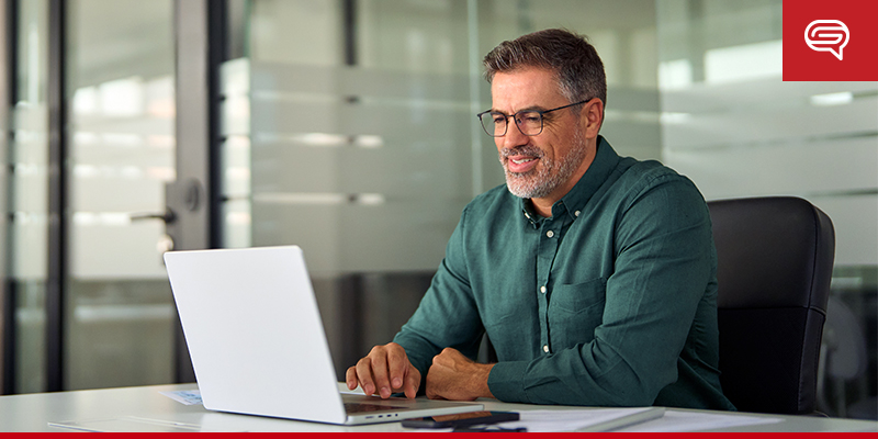 A man with glasses and a beard, wearing a green shirt, is sitting at a desk and working on a PowerPoint pitch deck in a modern office setting with glass partitions in the background. He appears to be focused and content.
