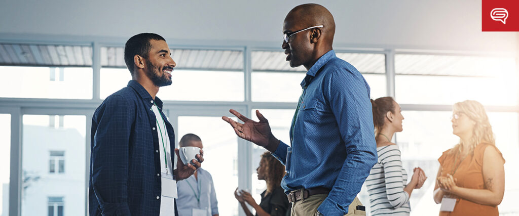 Two men are conversing with smiles, one holding a coffee cup, in a bright, modern office space. In the background, other people are mingling and chatting in small groups, suggesting a social or networking event. One man gestures towards his laptop screen, possibly discussing a pitch deck or presentation.