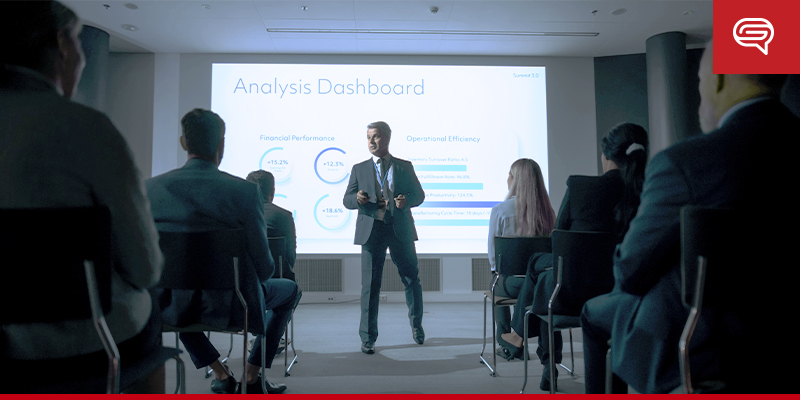 A man in a suit is giving a PowerPoint presentation in a dimly lit room. He stands in front of a large screen displaying an "Analysis Dashboard" with financial performance and operational efficiency data. Several people sit in rows, attentively listening to him.