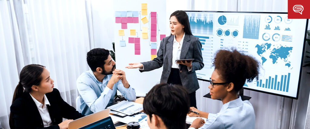 A woman in business attire is presenting a PowerPoint slide on a tablet to colleagues seated around a table. Behind her, a large screen displays various charts, graphics, and a world map. Sticky notes are arranged on a whiteboard in the background.