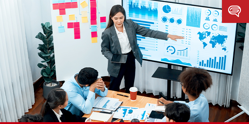 A woman stands and points to a large screen displaying various charts and graphs from her PowerPoint pitch deck in a meeting room. Five people sit around a table, looking at her presentation. There are sticky notes on a whiteboard and papers with data on the table.