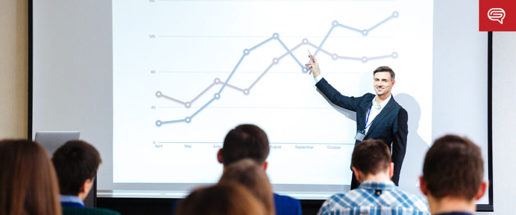 A man in a suit stands in front of a screen, pointing to a slide with a line graph showing two different-colored lines. He is presenting his pitch deck to an audience seated in rows, illustrating data trends over months labeled from April to October.