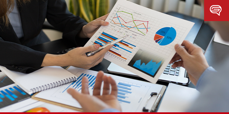 Two professionals sit at a desk analyzing various data charts and graphs on paper. One person points to a bar chart with a pen, while another holds up a slide displaying pie, bar, and line graphs. A notepad and laptop are visible on the desk, suggesting they are preparing for an upcoming presentation.