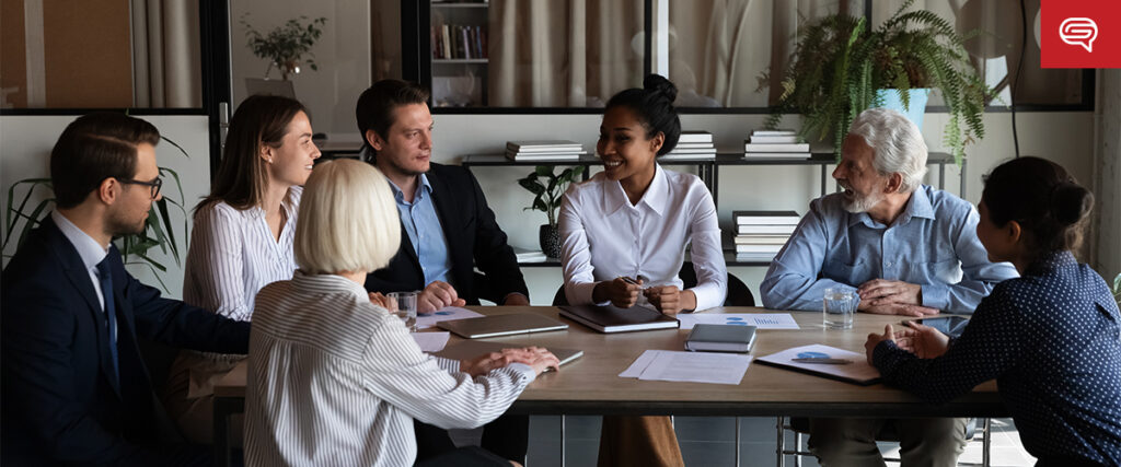 A group of six diverse individuals are seated around a table in an office, engaged in a meeting with slide templates visible on their laptops. Papers and notepads are on the table, and they are all dressed in business attire. Shelves and potted plants are visible in the background.