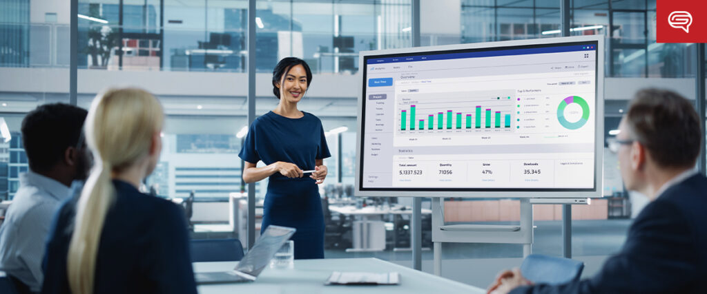 A woman stands in front of a large screen displaying PowerPoint slides with graphs and charts, presenting to a group of four colleagues seated around a conference table in a modern office. The presentation includes data visualizations, such as bar graphs and a pie chart.