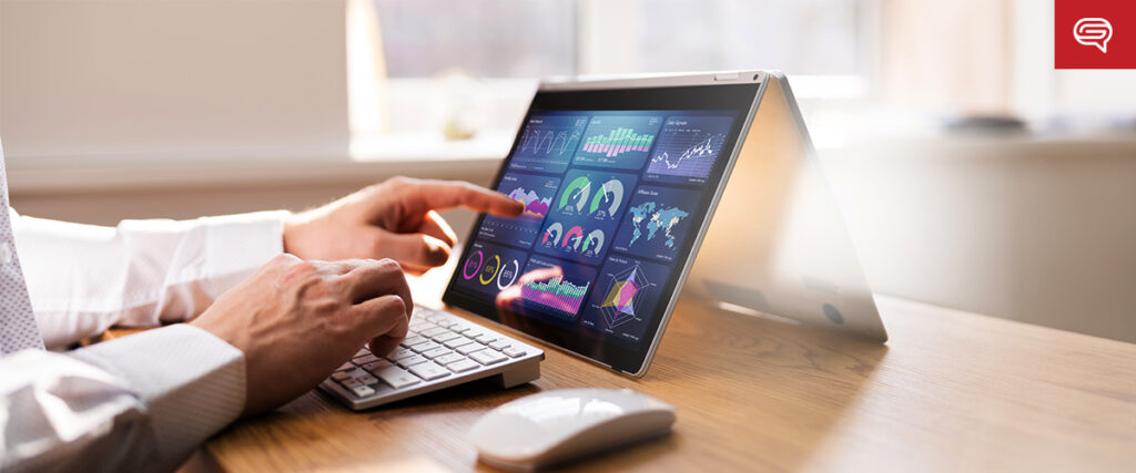 A person using a tablet in laptop mode on a wooden desk, interacting with colorful data visualizations on the screen. They are typing on an external keyboard and using a mouse while preparing their PowerPoint presentation. Sunlight streams through a window in the background.