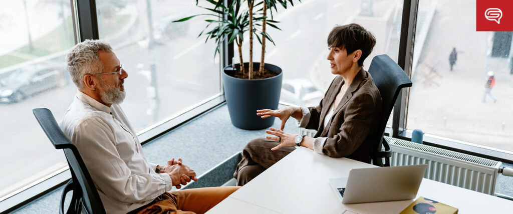 Two people are having a discussion in a modern office setting with large windows. One person, with gray hair and a beard, is showing a pitch deck on the laptop to the other, who has short, dark hair. They are seated across from each other at a desk beside a tall potted plant.