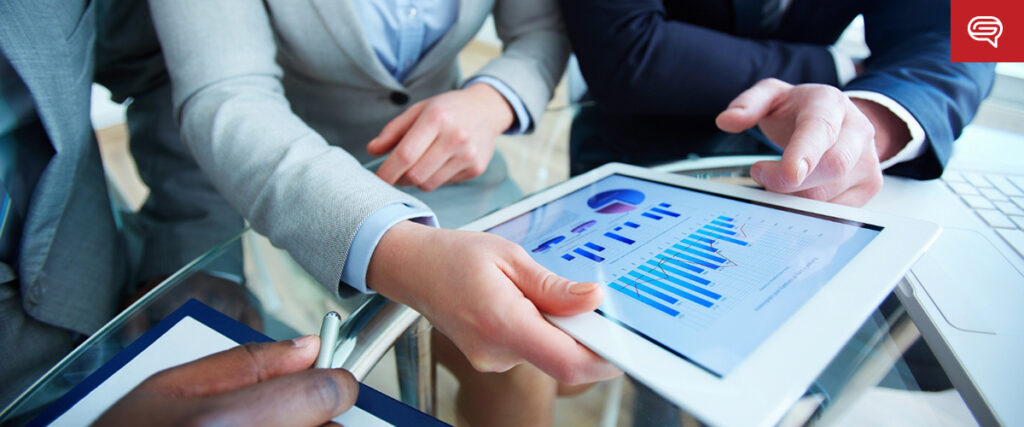 Three people dressed in business attire are gathered around a table, reviewing charts and graphs on a tablet. One person is holding the tablet, while another is pointing to the screen of a presentation. A laptop and documents are also visible on the glass table.
