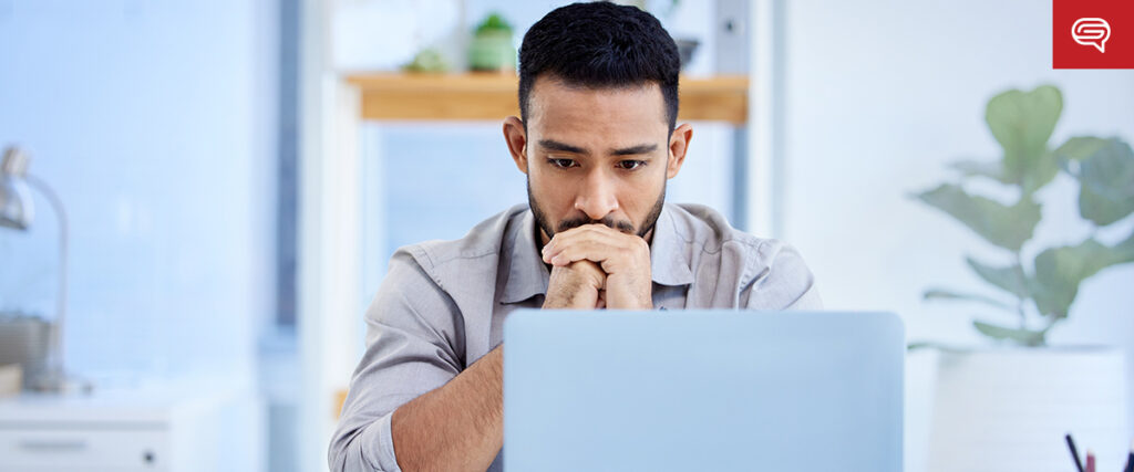 A man sits at a desk looking intently at a laptop screen with his hands clasped under his chin. The background features a blurred office setting with a potted plant and wooden shelves. A red speech bubble icon appears in the upper right corner, suggesting he is refining his pitch deck.