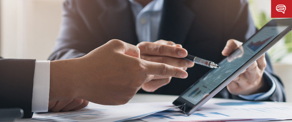 Two people in business attire discuss data displayed on a tablet, focusing on how to convert data sheets. One person points to the screen with a pen, while papers with graphs are visible on the table. The background is softly blurred, indicating a focused conversation.