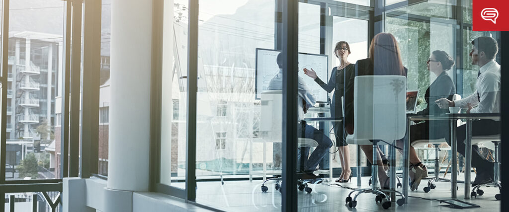 A group of people in a modern glass-walled office engage in a meeting. One person stands, presenting how to convert data sheets on a screen, while others sit around the table, listening attentively. City buildings are visible through the windows.
