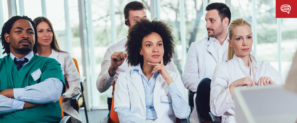 A group of medical professionals, including doctors and nurses, are seated in a classroom setting attentively listening to a presentation. They are wearing scrubs and white lab coats. The room is bright with large windows in the background, enhancing the clarity of the slide template being discussed.
