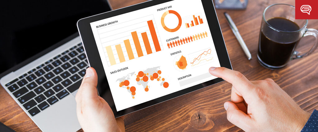 A person holding a tablet displaying various business analytics, including bar and pie charts, a world sales location map, and statistical graphs. A laptop showing converted data sheets, a notebook, a pen, and a cup of coffee are on the wooden desk in the background.