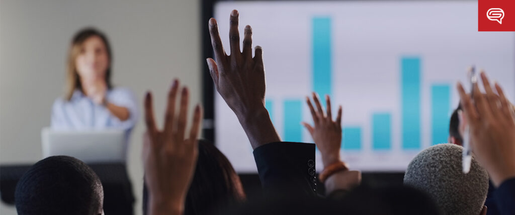 People raising their hands in a seminar or conference room, eager to ask the presenter about converting data sheets, as they stand by a screen displaying a bar chart.