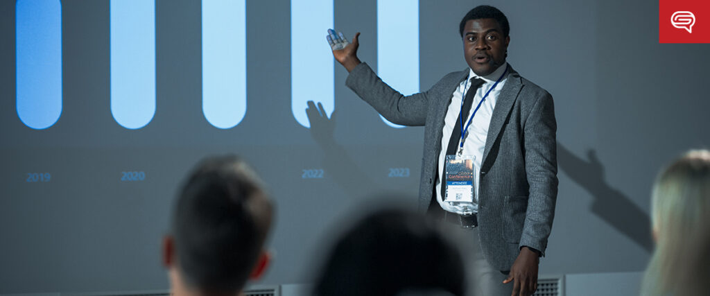 A man in a suit is giving a presentation, flipping through a digital chart like a flipbook. The projected chart displays bars labeled with years from 2019 to 2023. Audience members are listening attentively in the foreground.