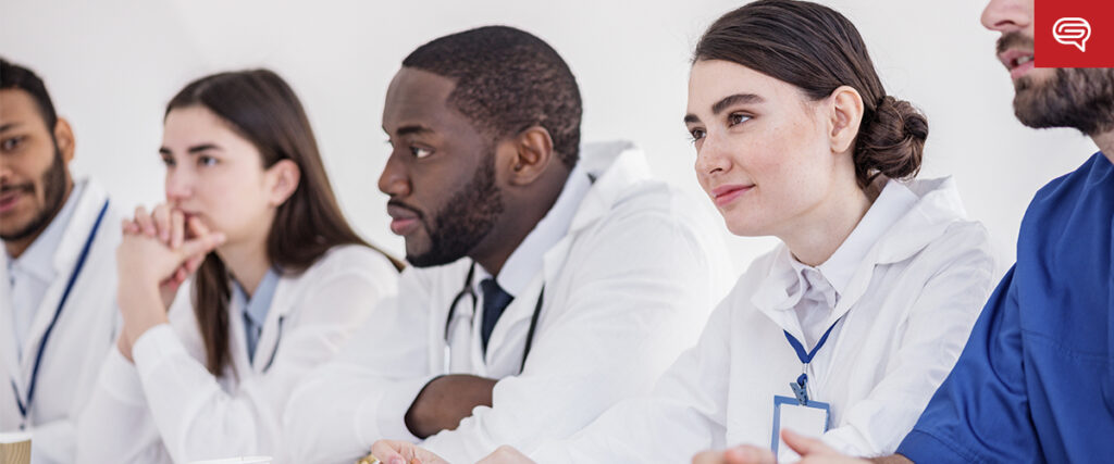 A group of medical professionals in white coats sits in a row, attentively listening to life science presentations. They are diverse in appearance and wear ID badges, suggesting a seminar or meeting environment.