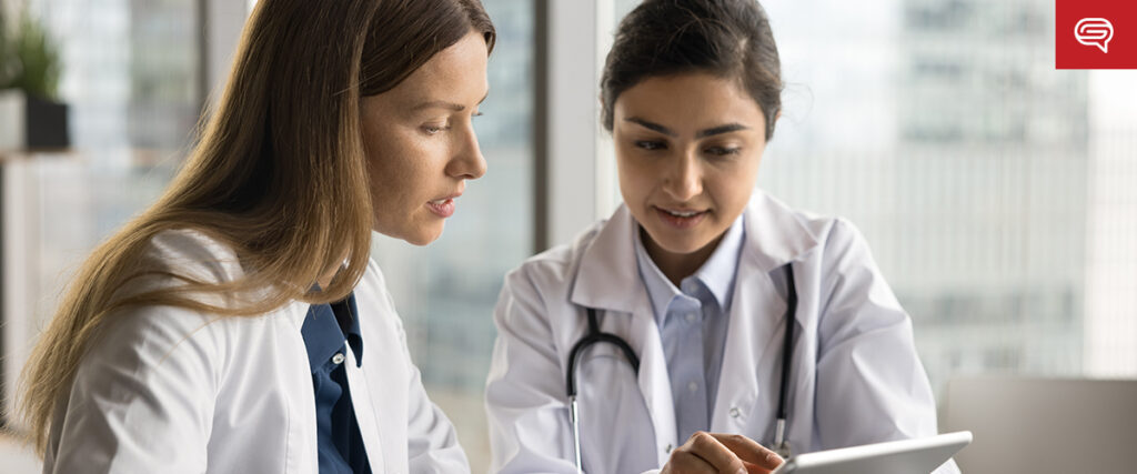 Two doctors in white coats are flipping through a digital flipbook on a tablet. The woman on the left has long brown hair, while the woman on the right, with dark hair tied back and wearing a stethoscope, remains focused and engaged.